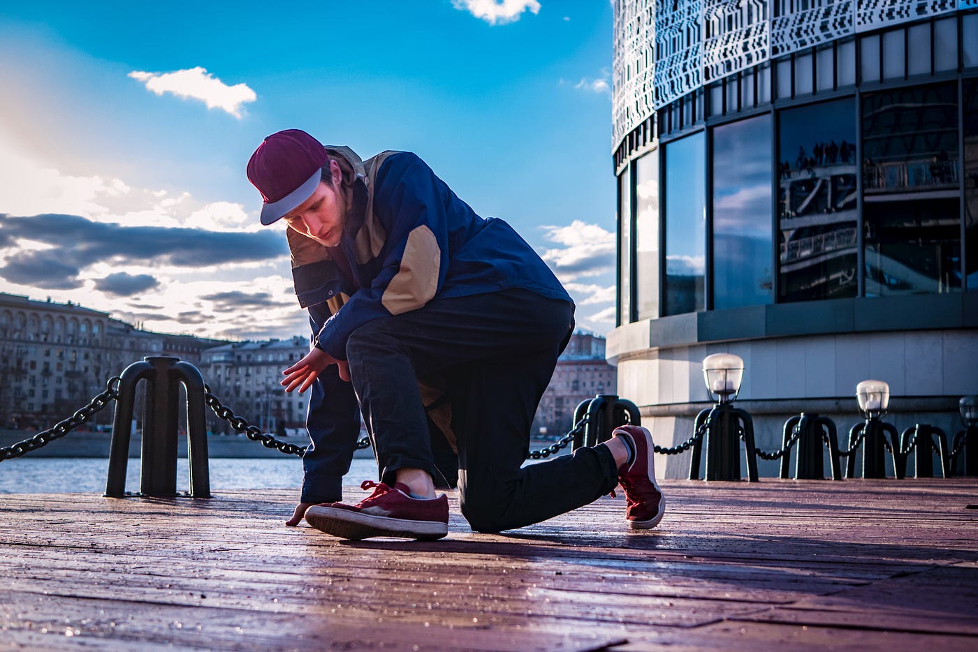 A man break dances on the damp wood floor of a pier, buildings and a susnet in the backgroud.