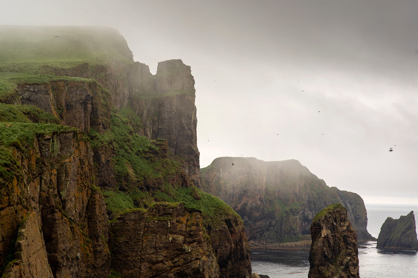 Green and rocky cliffs along a coastline with bands of fog and clouds.