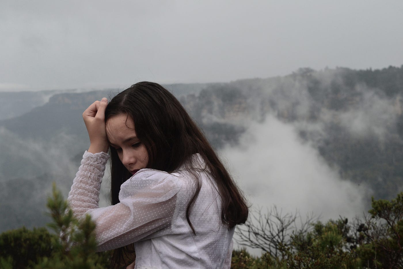 a young woman with long dark brown hair, looking sad with a white shirt, holding up her arm with her head tucked into her arm, with a cloudy background and some bits of dark green foliage in the foreground