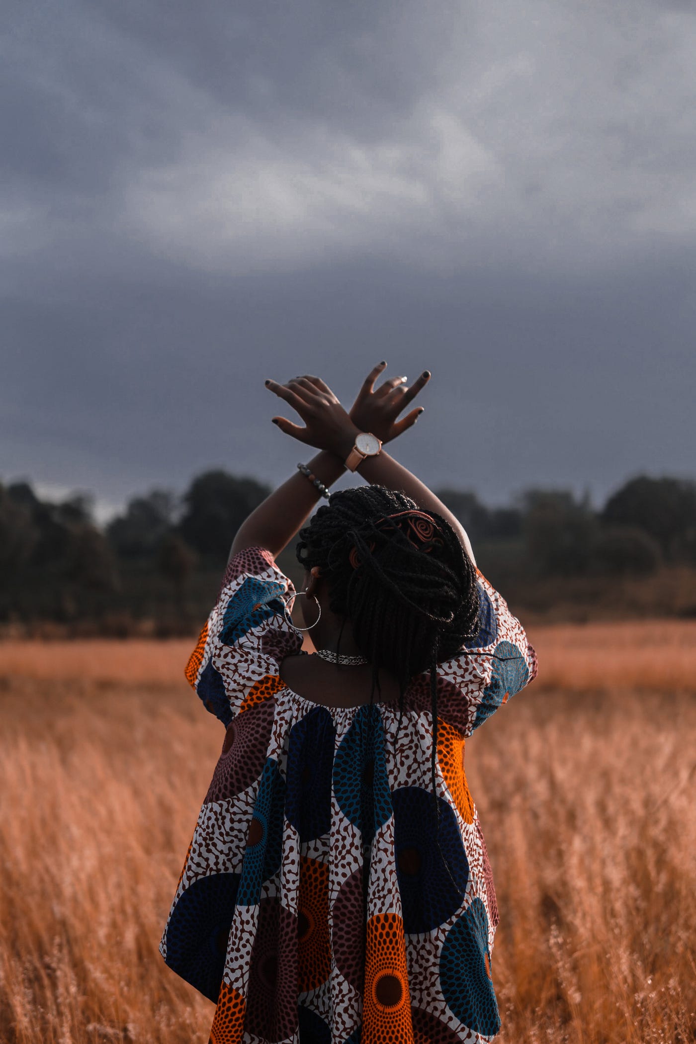 Black womxn with braids in African print dress, back to camera, hands up towards a grey sky.