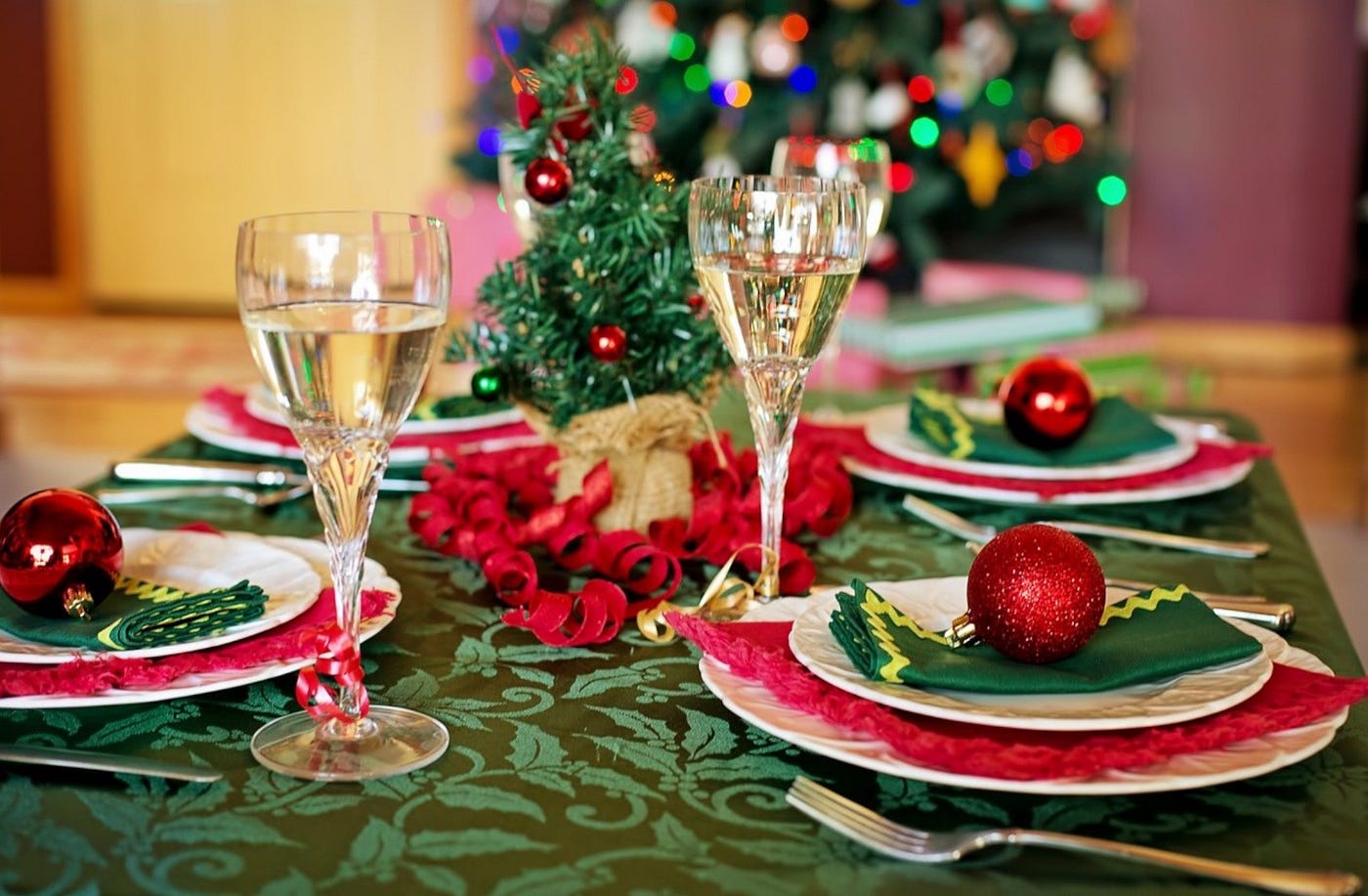 Color photo of a table decorated for a Christmas or winter holiday dinner. A green tablecloth covers the table with filled champagne glasses, silverware, plates set and ready with a partial view of a Christmas tree.
