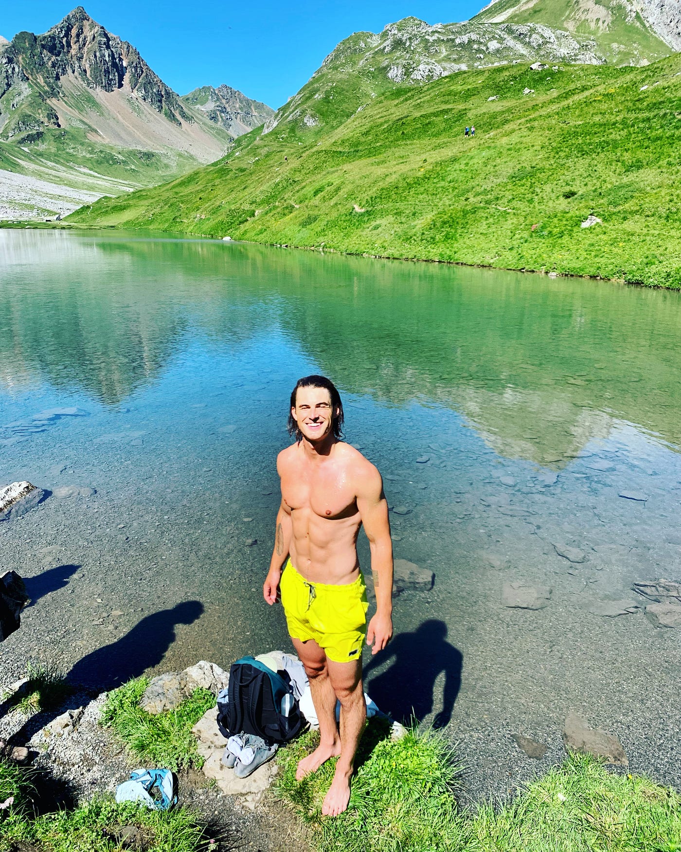 A fit man in his late twenties, standing in wet bathing trunks on the shore of a frigid mountain lake in Arosa, Switzerland.