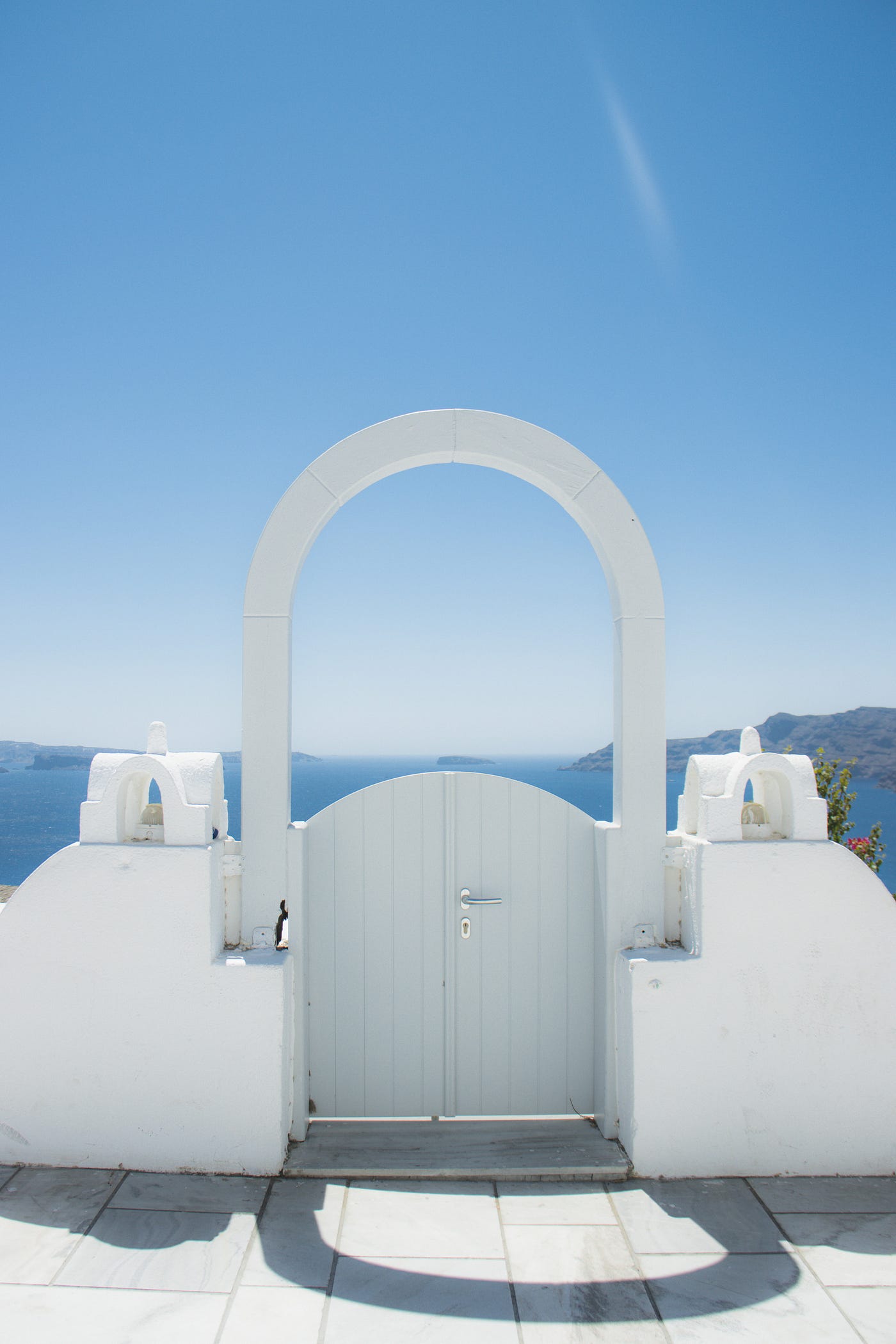 The image shows a gate with an arc in Oia, Greece. In the background you will find blue sky and a view of the sea.
