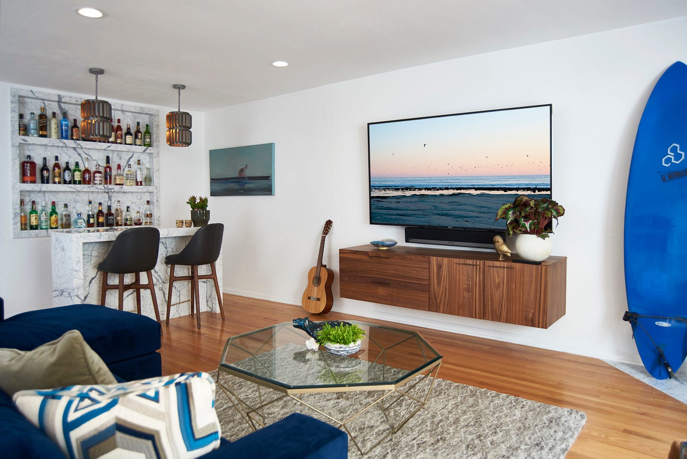A living room with a blue couch, glass coffee table, and beige rug. On the wall facing the couch, there is a wall mounted television with a wall mounted media console, a blue surfboard and a guitar. There is also a home bar visible in the background.
