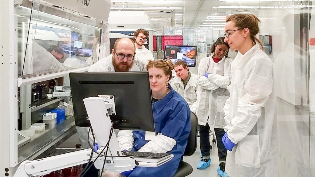Six scientists in white lab coats huddle around a computer screen to examine cells generated by the NYSCF Global Stem Cell Array®