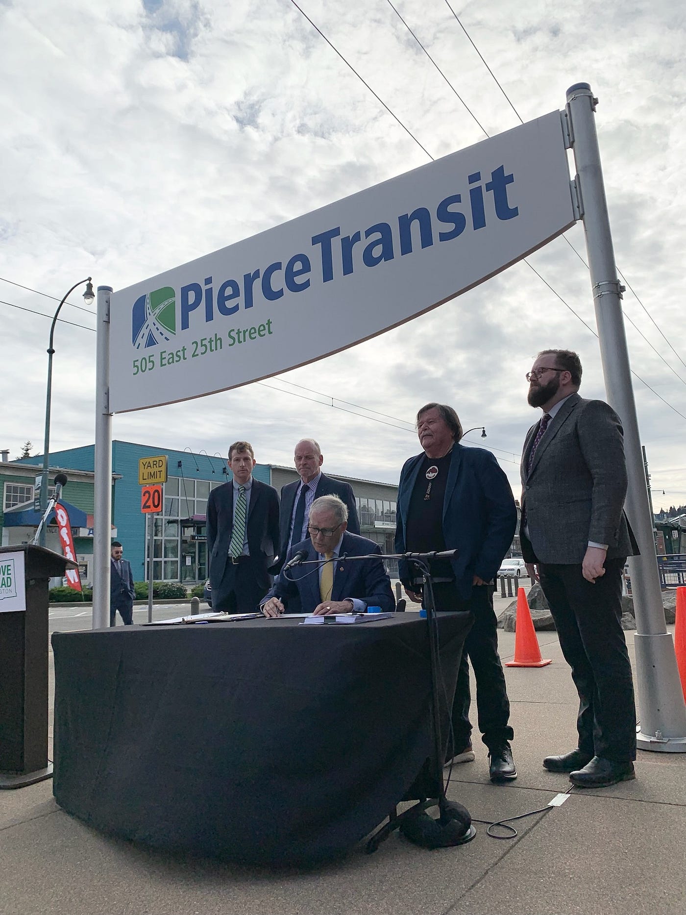 The governor is seated at a table outdoors signing a bill. Behind him are four men who are watching and smiling. They are under a large Pierce Transit sign.
