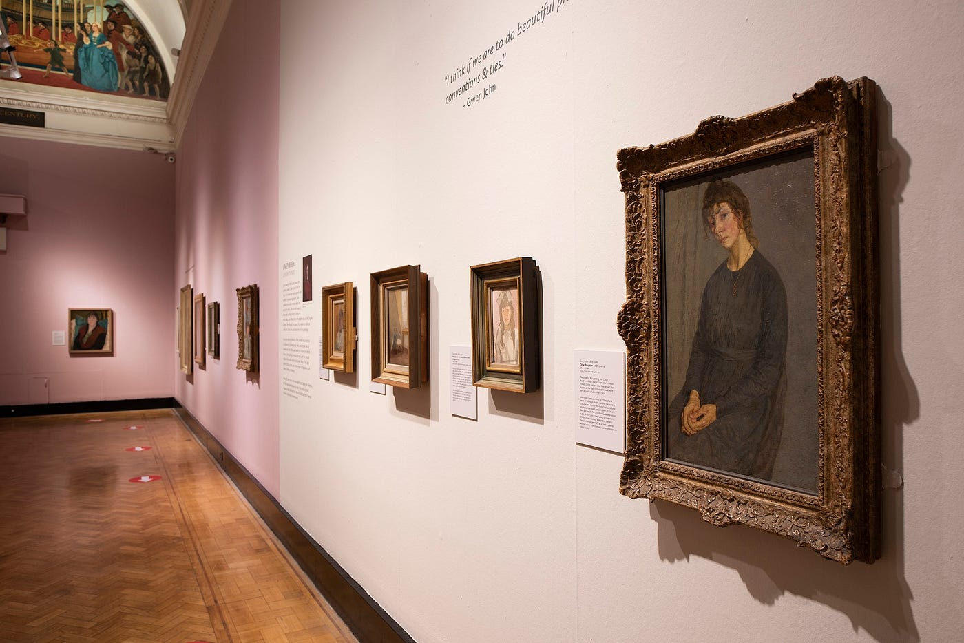 A photo of a gallery wall with gold frames hung on it, with a painting of a sitting woman wearing a black dress