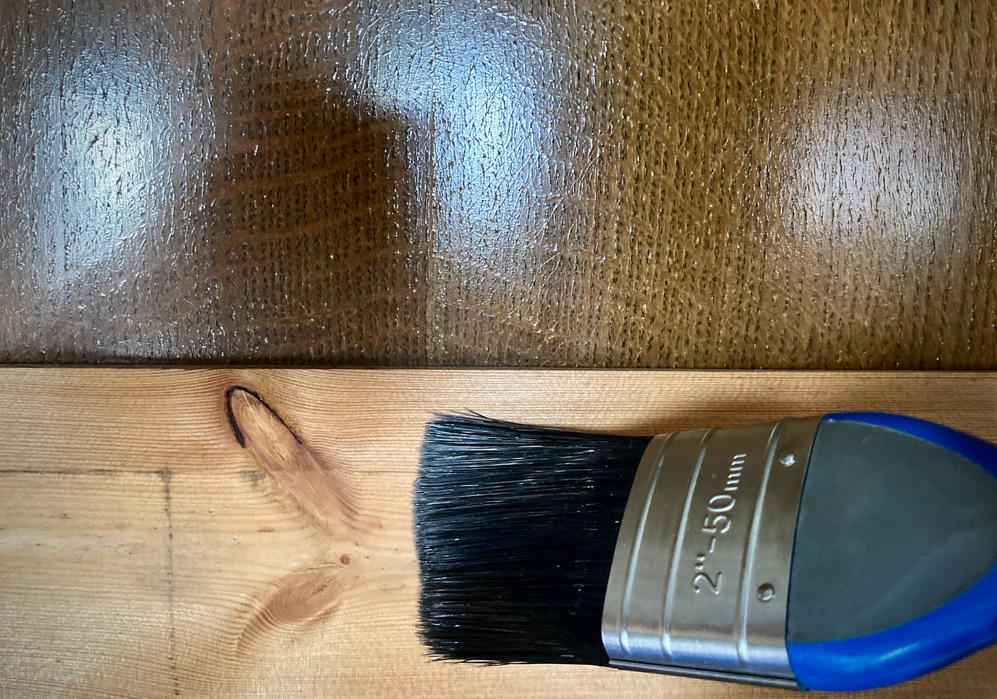 A paint brush beld against some bare skirting board. The background is a dark shiny wooden surface.
