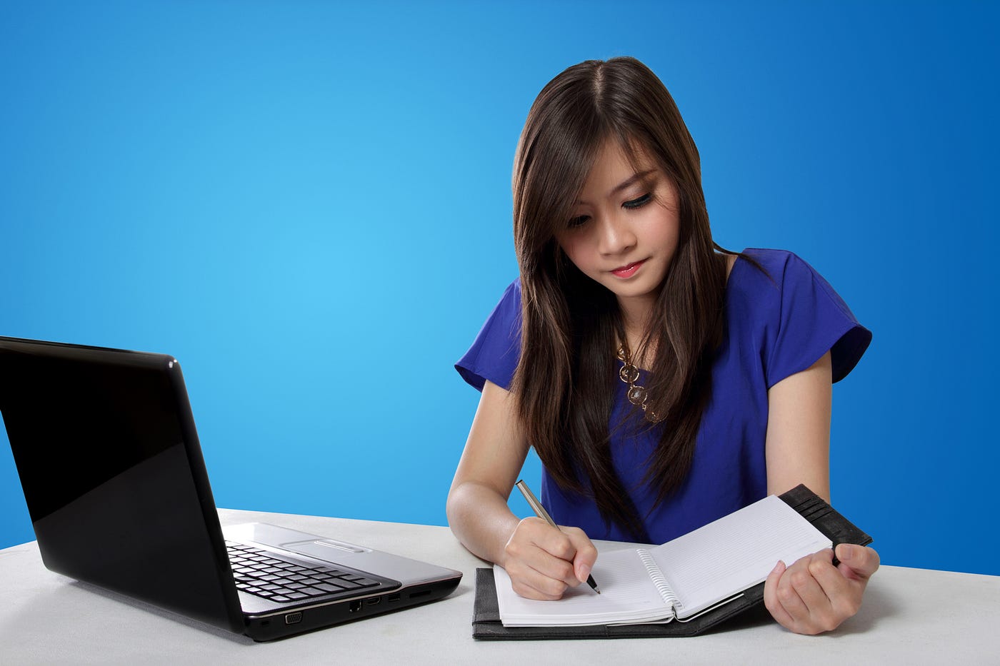 Beautiful Asian girl with long hair writing in a notebook. An open laptop sits next to her at a white table. The background is blue.