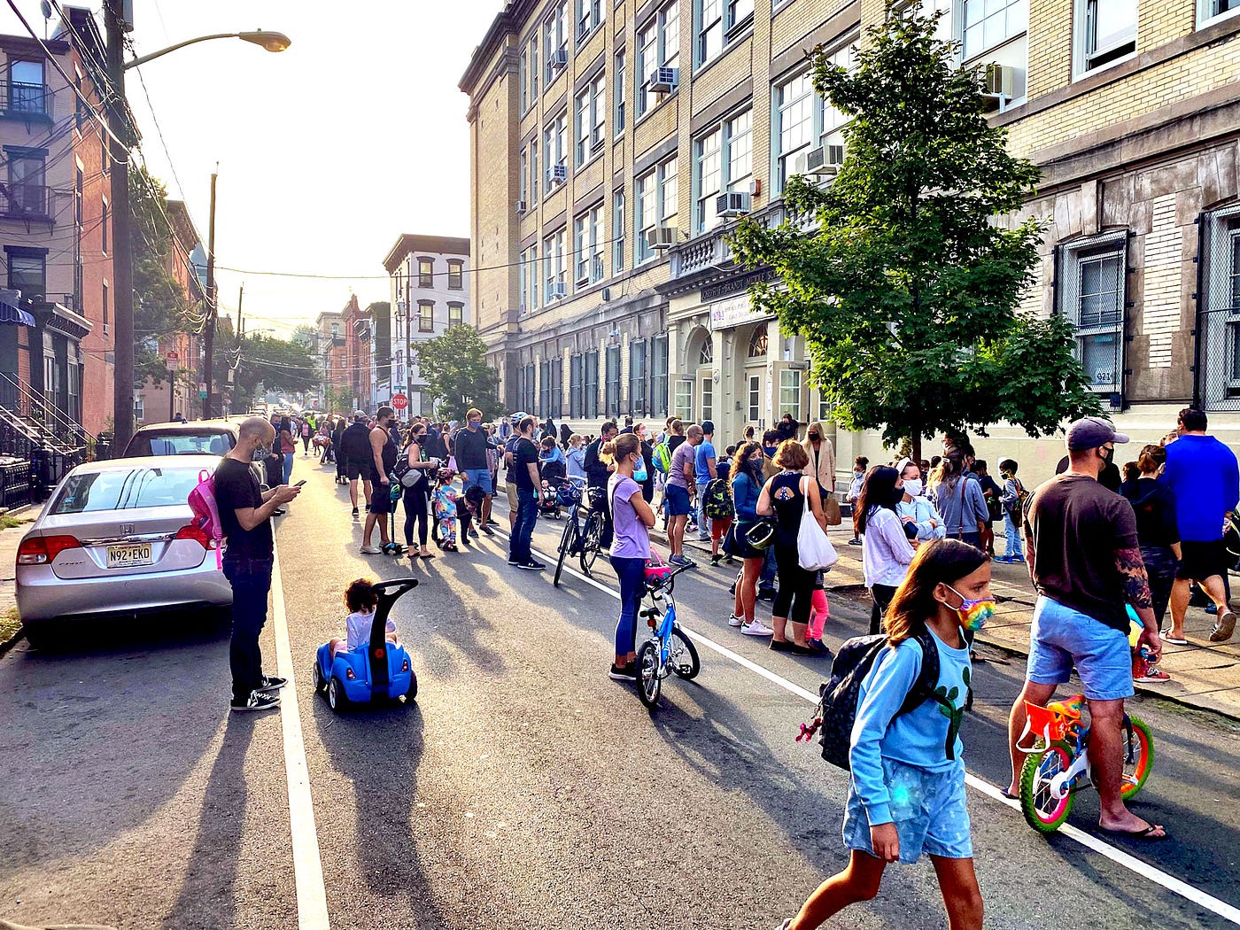 Children and parents gather in a street in front of a school with late afternoon sun slanting through and casting long shadows.