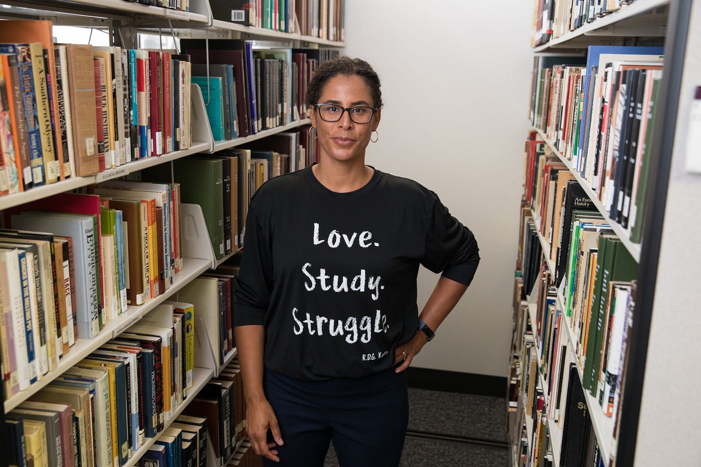 woman with black shirt standing in stack of books at library. shirt says: Love, study, struggle