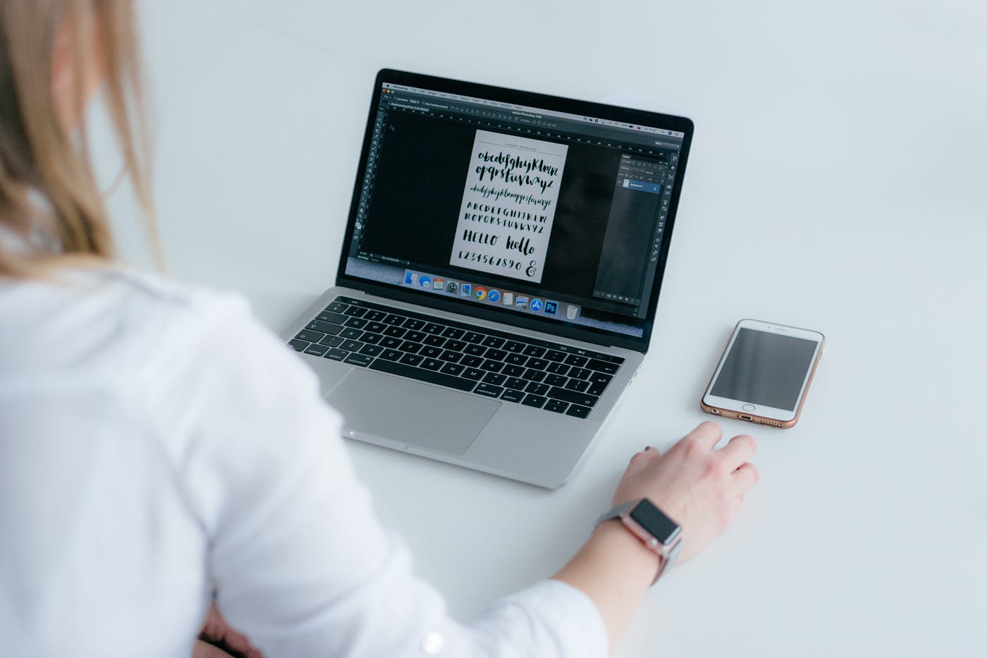 Photo of a woman with her back to the camera looking a laptop screen. On the screen a design program that looks like photoshop is open, and next to the laptop is a cell phone.