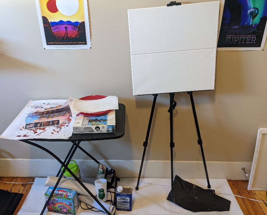 Photo by the author of her art space that she has needed to take a break from based on acquiring supplies, life, and more. A table on the left has half finished, unstarted art projects, and supplies waiting to be used both on top and beneath it. On the right, 2 canvases sit atop an easel, waiting to be used.