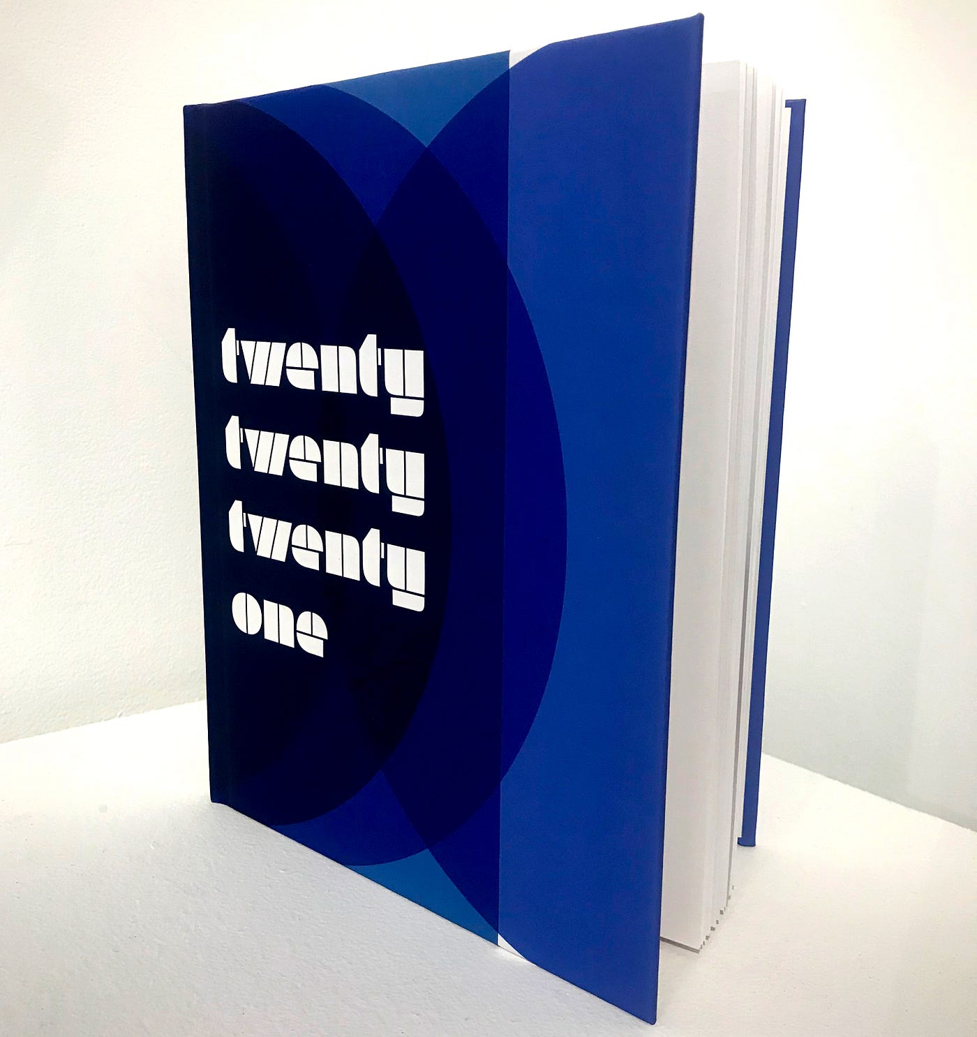 A book, resting upright and slightly open, rests on a white surface witha white background. The book’s cover has blue circular forms in an abstract composition, with the title “202021” in white typography.