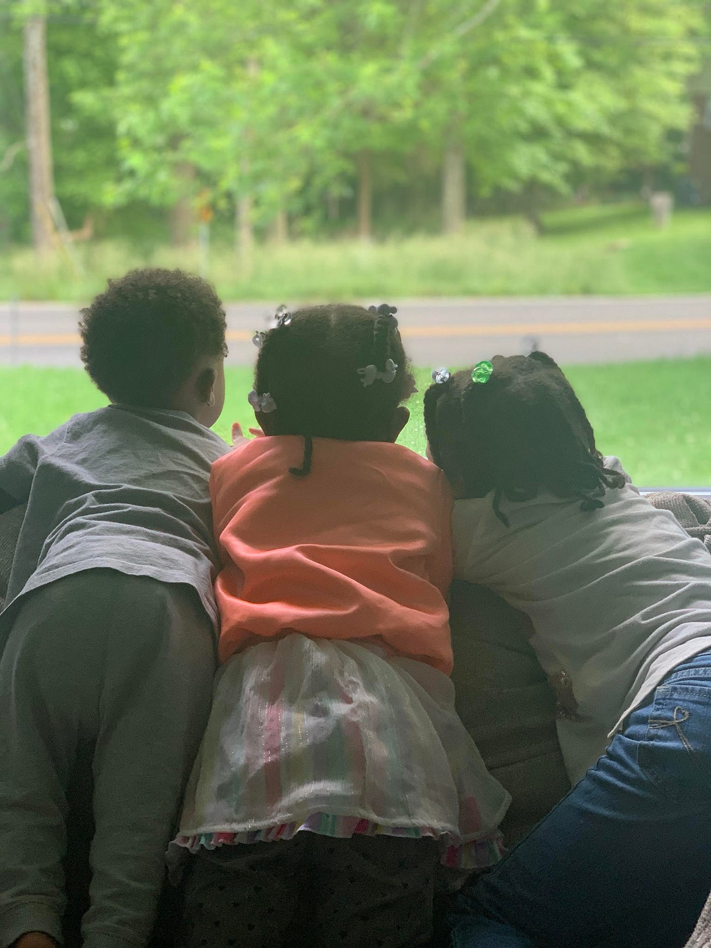 3 young Black children seen from behind, looking out the window at grass and a road.