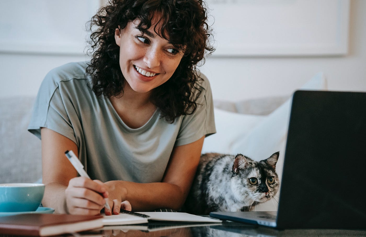 Smiling woman looking at computer screen, taking notes with cat next to her. Sandra Pawula 2021 Medium Library