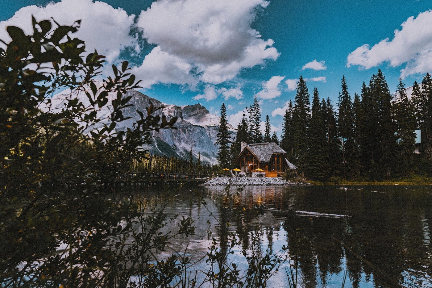 Color photo of a lake side cabin. The day is blue sky, few scattered clouds, trees.