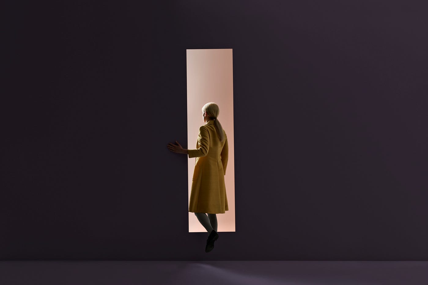 A woman in a dark room stepping through a rectangular cutout in the wall and into a brighter room.