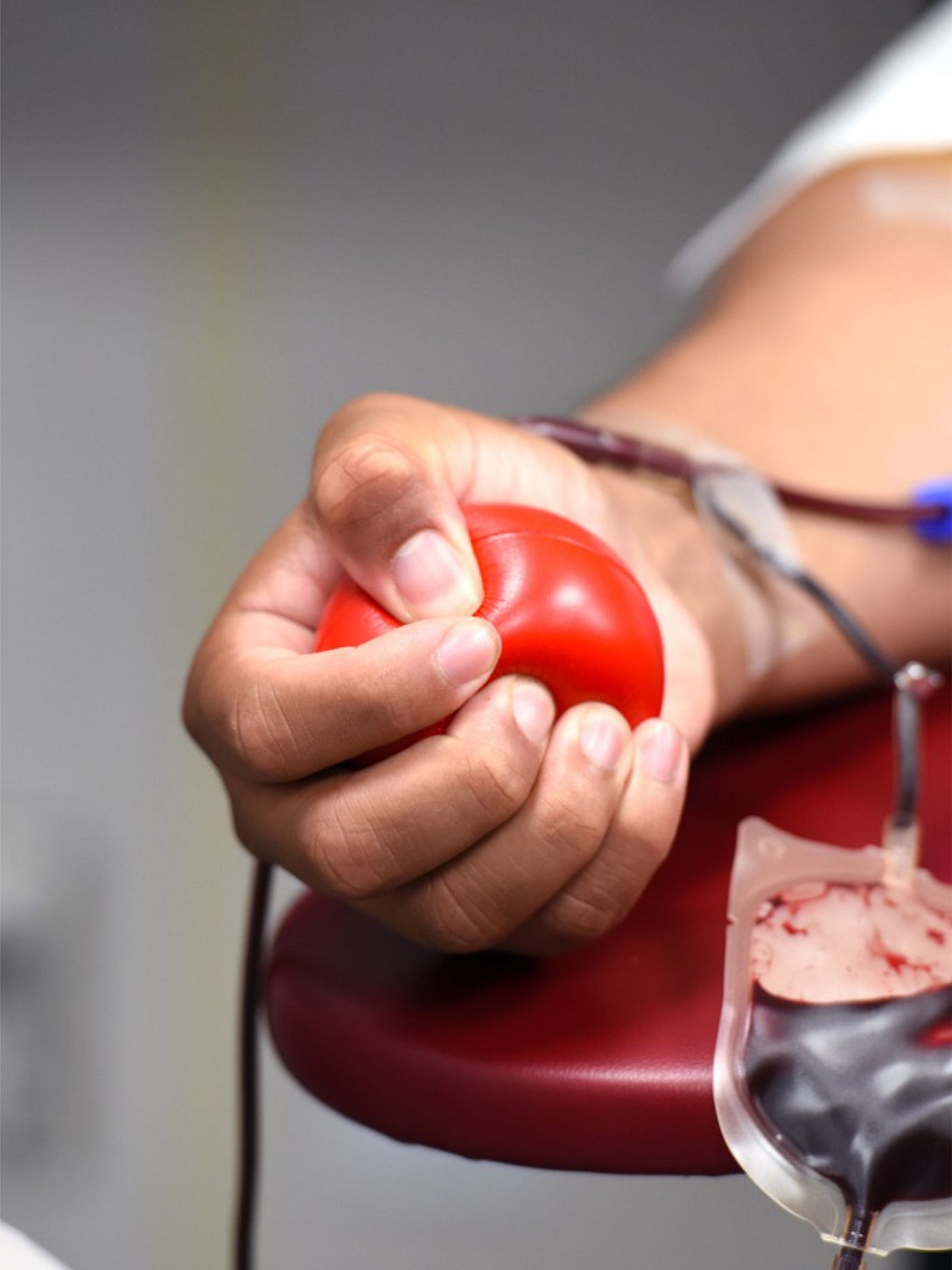 Color photo of an individual squeezing a red rubber ball in their hand, while they donate blood.