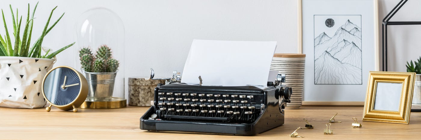 A black typewriter sits on a desktop with stationary and pictures around it. A blank page sits in the typewriter just waiting for inspiration