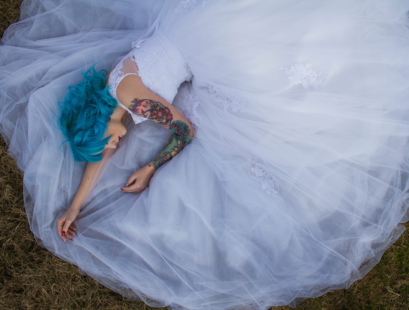 Woman with blue hair and tattoos in white wedding dress laying on the ground.