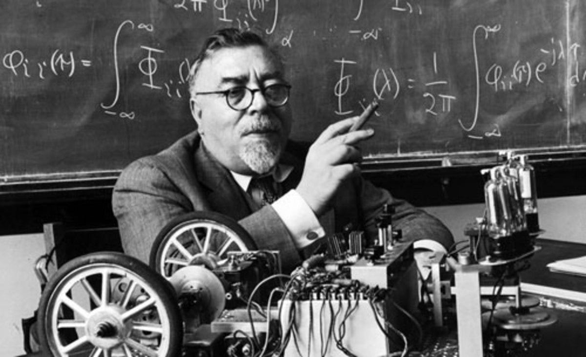 Cyberneticist Norbert Wiener sitting in front of a blackboard with the wheeled robot, Palomilla