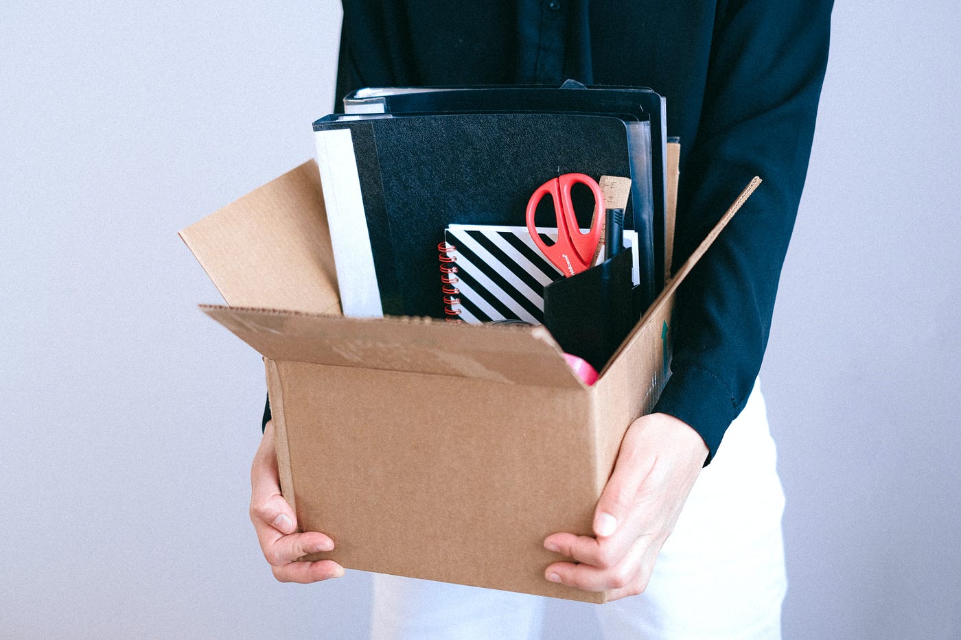 Torso of a female in white slacks and a navy shirt holding a cardboard box full of office supplies
