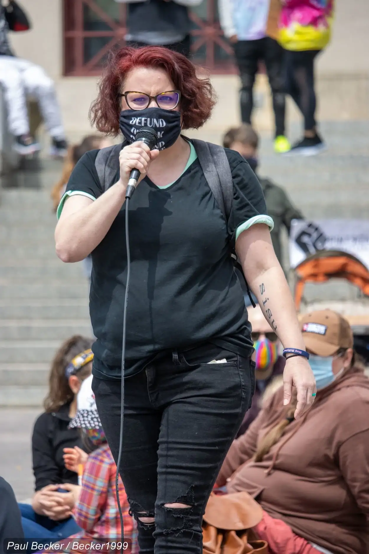 a photo of me, a white woman wearing a black tee and black pants, a black mask that says “disarm, disband, defund the police” with the words somewhat obscured. my hair is short, just to the ears, darker red, and I’m wearing black rimmed glasses, somewhat scowly. There are people behind me on steps in the background and a BLM fist on a sign.