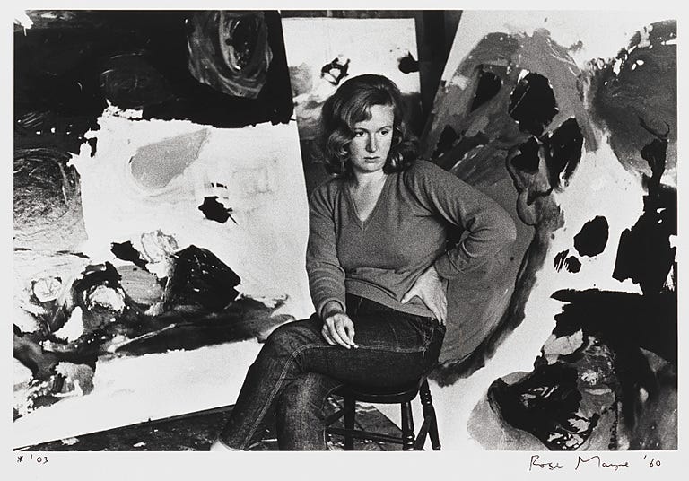 Photograph of woman with brown hair, jumper and jeans sitting in front of art