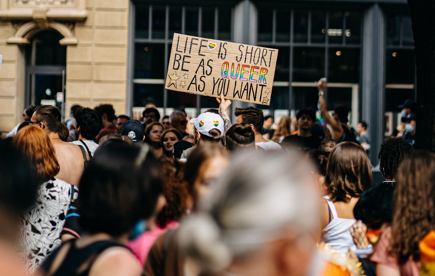 Part of the crowd at a Pride Parade. In the center is a person holding up a sign, which says “Life is short be as queer as you want” in all caps. The word queer is in the colors of the rainbow. The person standing next to them is wearing a white baseball cap with rainbow hearts on it.