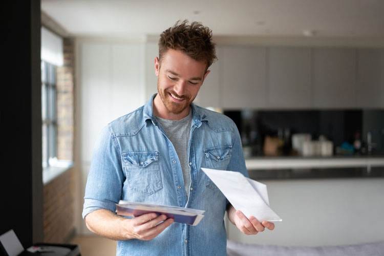A man is standing and looking at envelopes he holds in both hands separately.