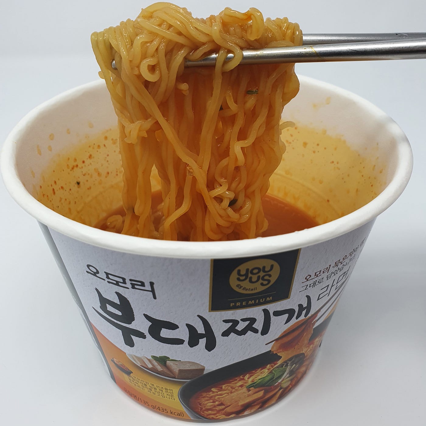 A finished cup of YouUs Omori Budae-jjigae Ramen still in the cup with chopsticks holding up the noodles.