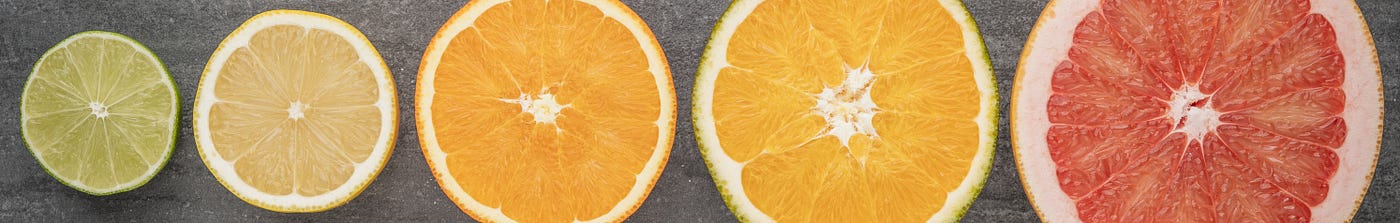 Five cross sections of citrus fruits arranged in ascending order of diameter on a textured grey background. From left to right, they appear to be a lime, a lemon, and orange, a larger orange citrus fruit, and a red grapefruit.
