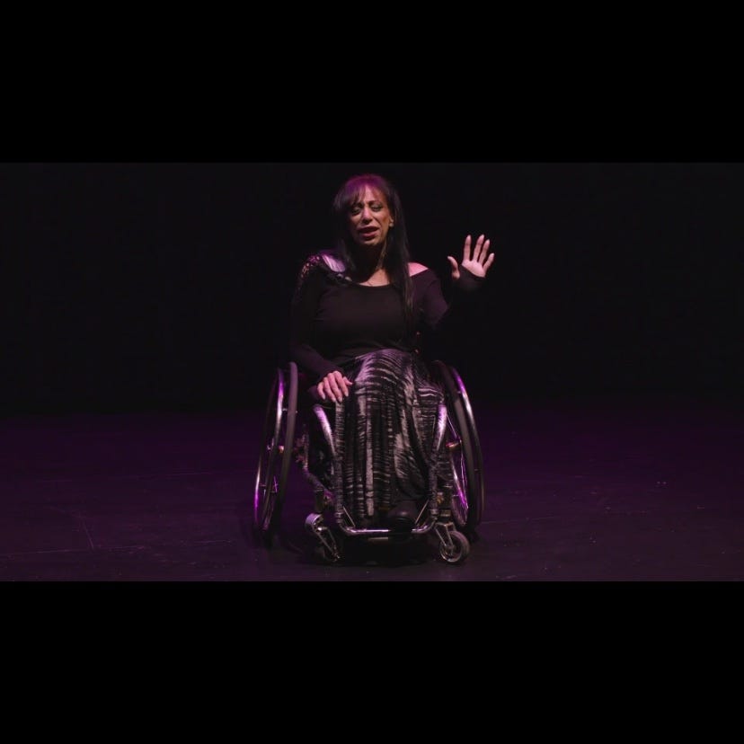 a Latina woman in her wheelchair, wearing all black, with her left hand raised palm toward the audience