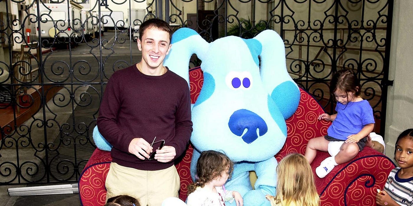 Steve Burns in a purple sweater and khaki pants with a Blue face character sitting in a thinking chair, surrounded by toddlers.