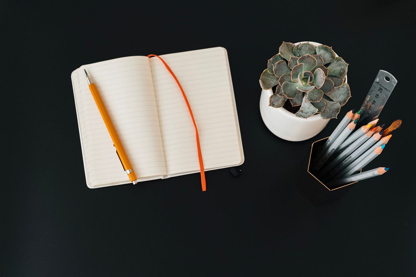 An open notebook and yellow pencil on a black background. There is also a small potted plant and a pencil stand with a bunch of pencils in it.