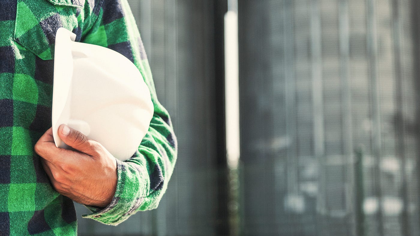 A construction worker in a green plaid shirt is holding a white hard hat against a gray background.