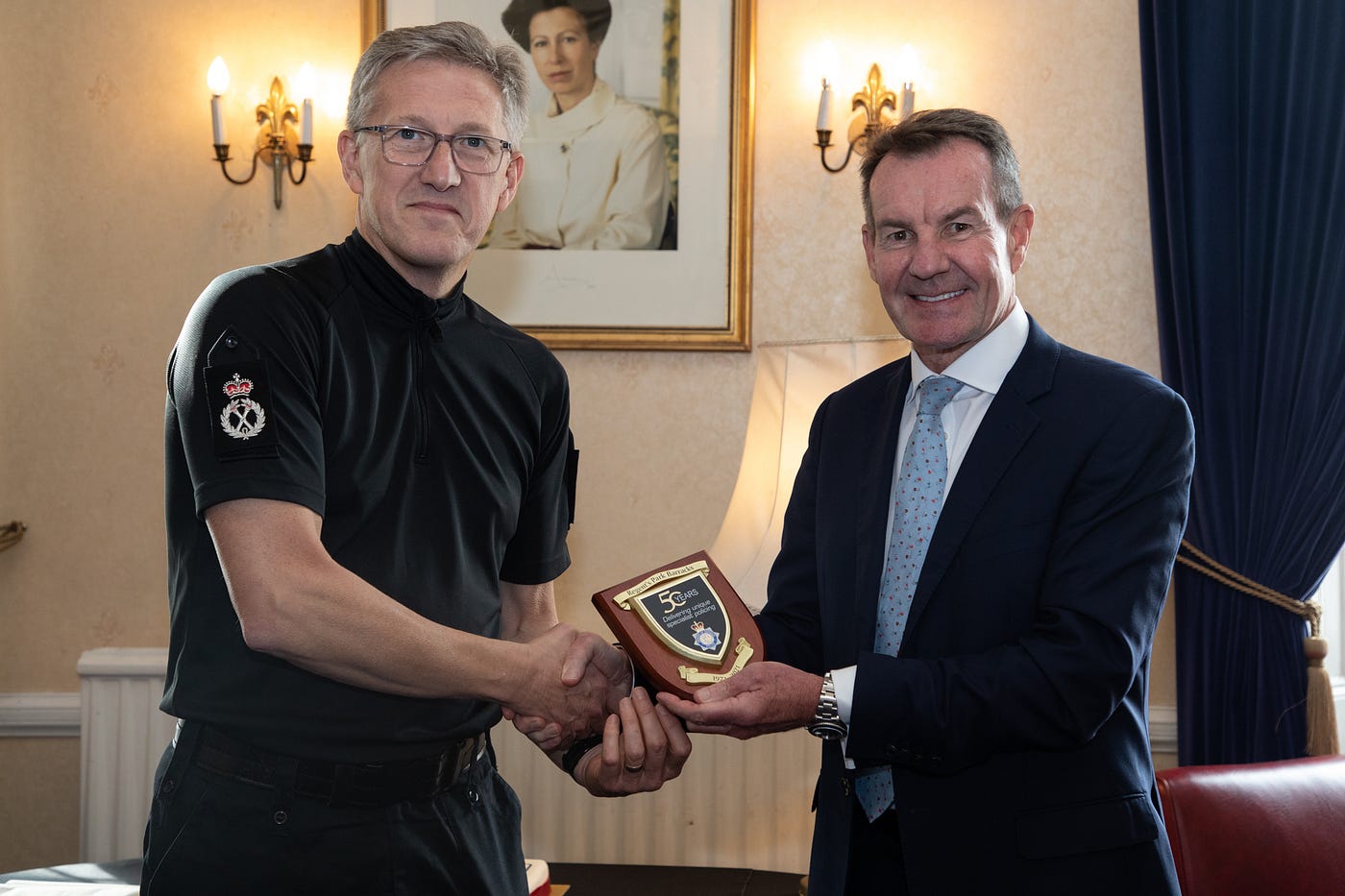 Chief Constable Andy Adams handing Defence Chief Operating Officer Mike Baker a MDP plaque, wall lights, and picture of Queen Elizabeth II on the wall in the background