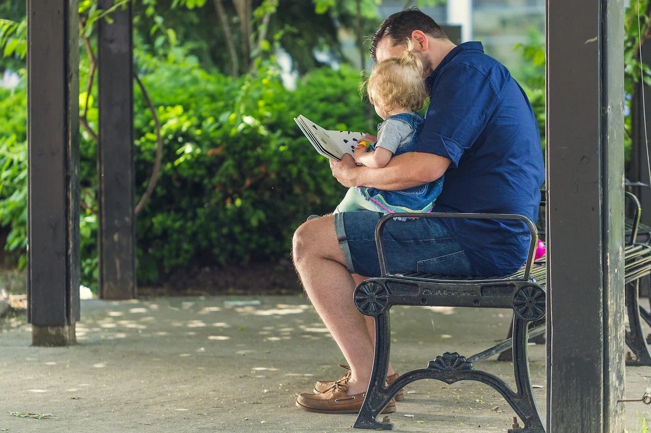 An image of a father reading a book to a child on his lap on a bench.