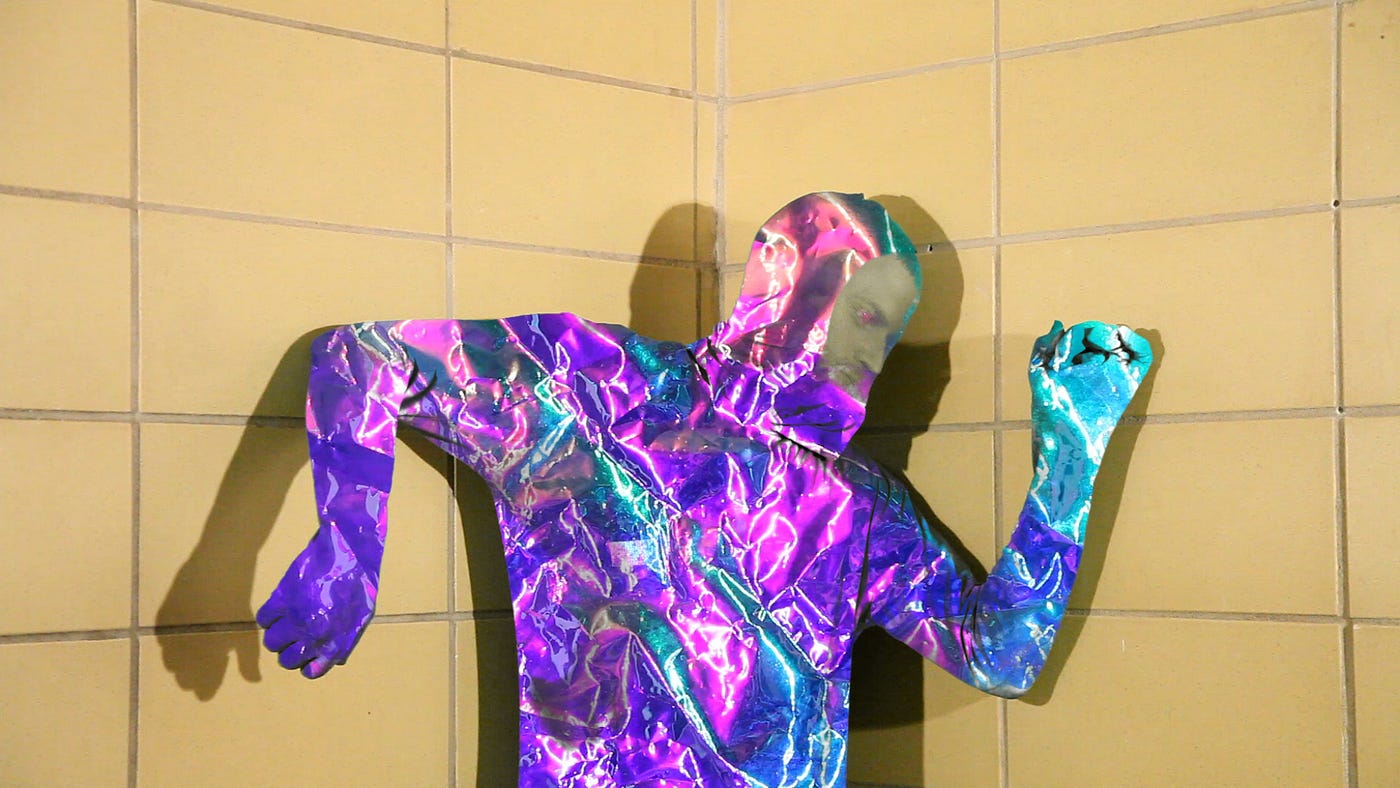 Photograph of the author in front of a khaki-colored tile wall wearing a iridescent mylar suit.