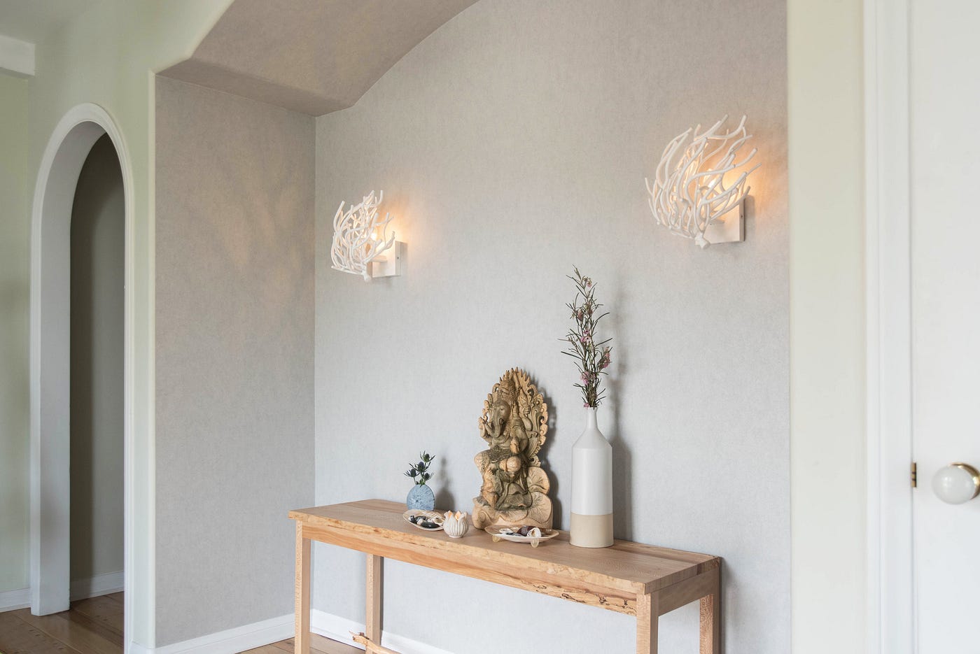 A welcoming foyer with plants and art pieces inspired by nature, like these delicate coral inspired wall sconces. There is a wooden table holding a display of vases, a figurine, and small natural objects.