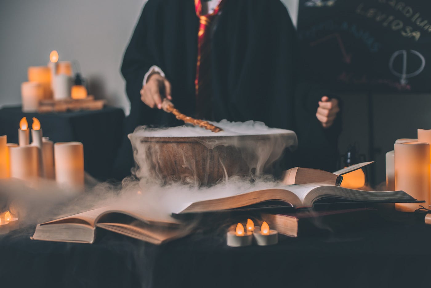 Harry Potter-like setting wtih open books, lit candles and someone in wizards clothing waving a wand over a smoking kettle