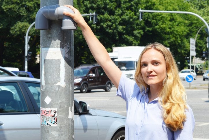 A person posing next to an air quality sensor on a pole next to a busy intersection in an urban setting/