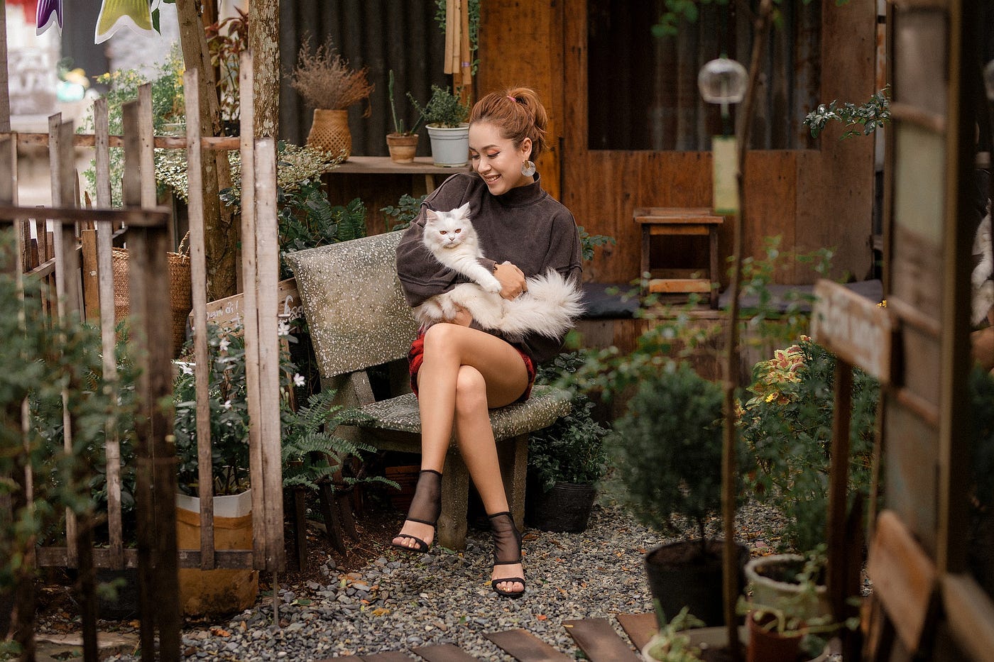 An Asian woman on an outdoor garden bench holding and smiling at a fluffy white cat.