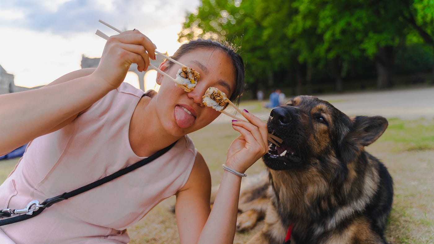 a girl on beach holding food in chopsticks over each eye her tongue is sticking out her dog trying to bite nip the end of a chopstick