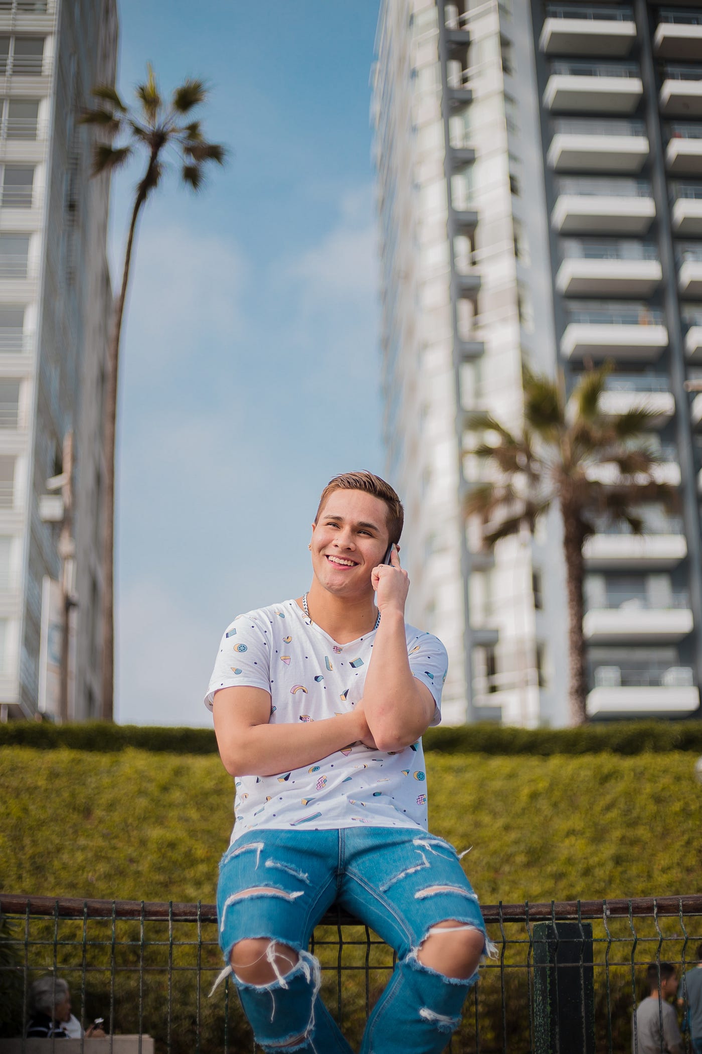 A man is talking on the phone and he’s wearing a white short sleeved shirt and jeans. There are buildings and palm trees behind him. He is sitting outside.
