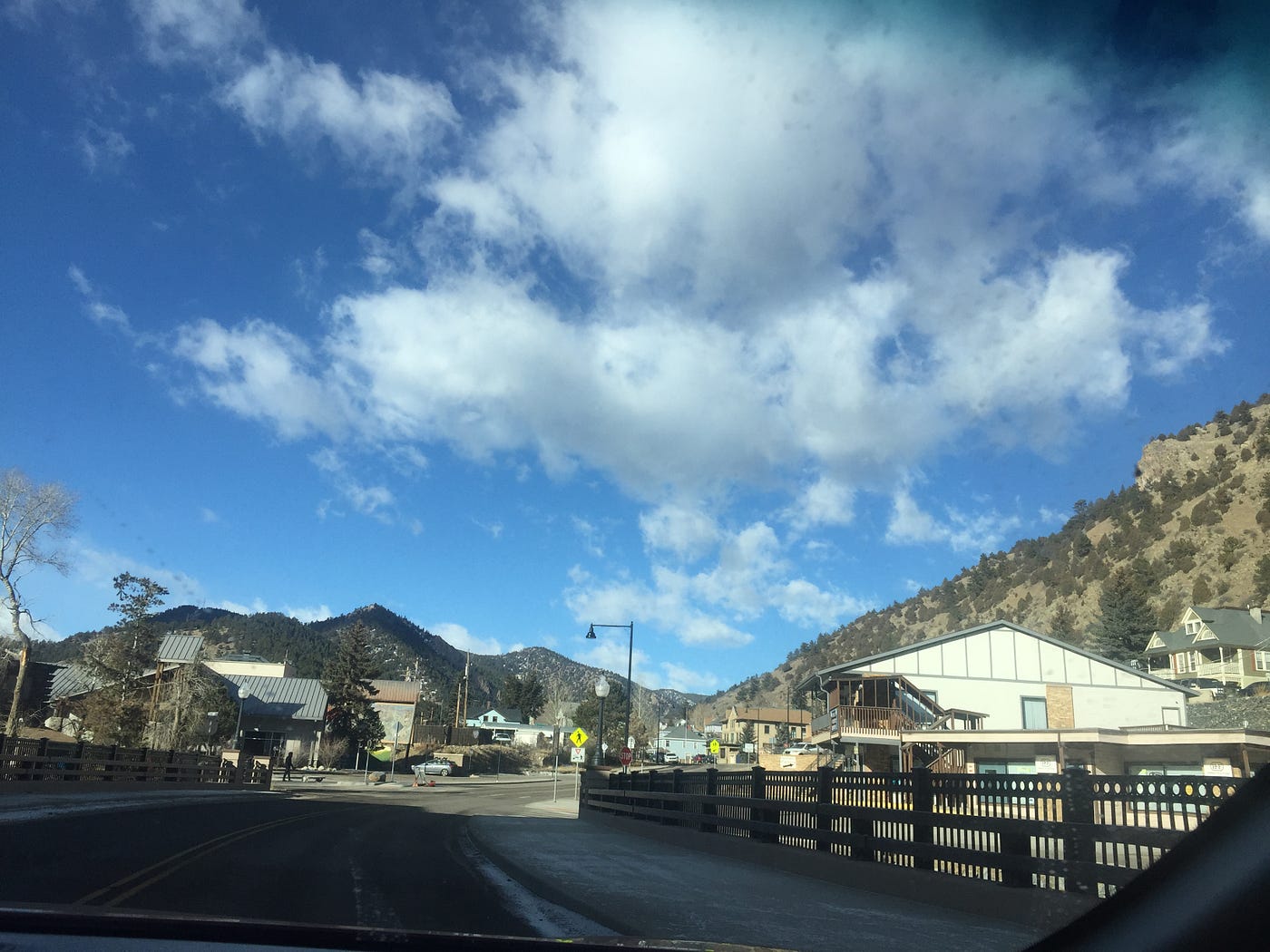 Deep blue, cloudy sky set over the mountains of Idaho Springs. Just below the mountain line is a small town with older buildings. Point of view: driving into Idaho Springs.