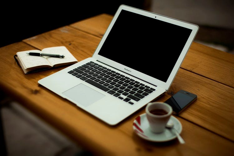 An image of a laptop, a notepad and pen and a cup of coffee on a table.