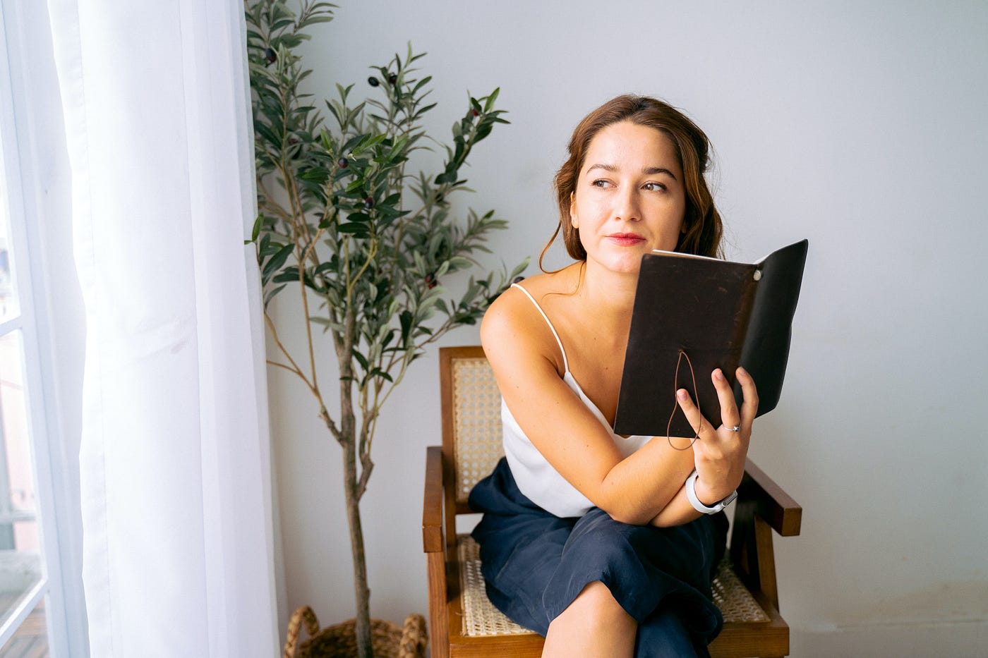 Girl sitting on brown chair holding a black notebook, staring into the distance