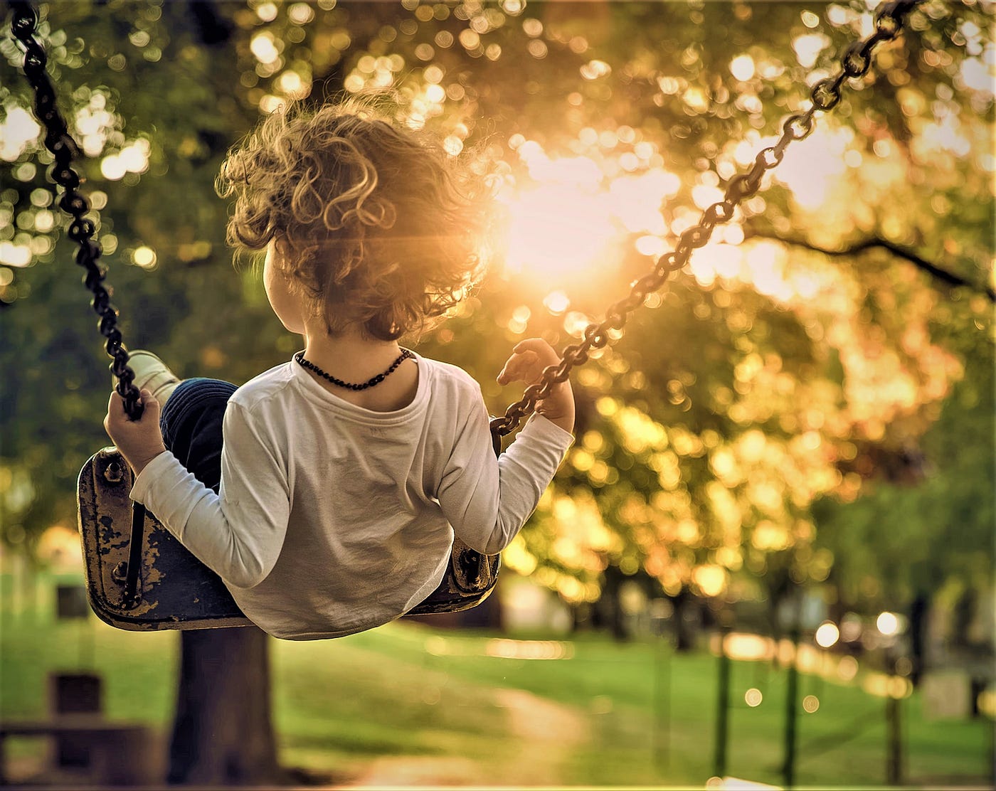 rear view of young boy with curly brown hair wearing long sleeve white top mid-air on a swing over grassy lawn